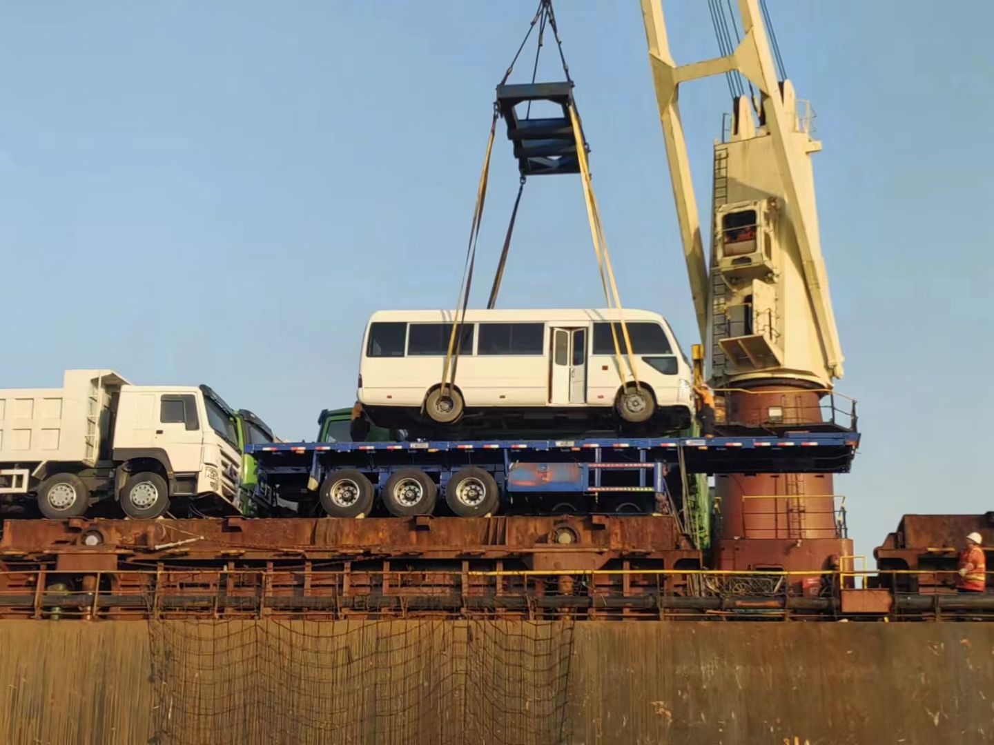 A coach bus lifted by ship crane onto a bulk carrier, dump trucks lined up on deck