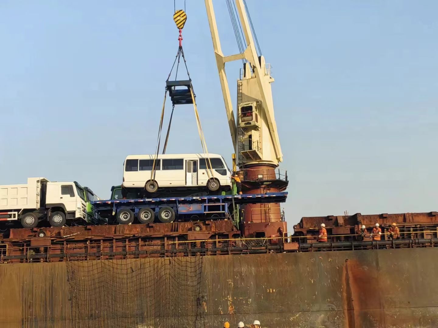 A coach bus raised on lifting slings above a flatbed trailer on the vessel deck