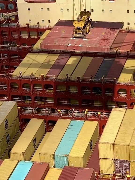 A quay crane lowering a flat rack onto a container ship between stacks of boxes