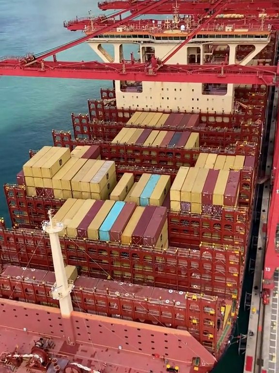 Aerial view along a container ship deck under red gantry cranes, with more cranes on the far shore