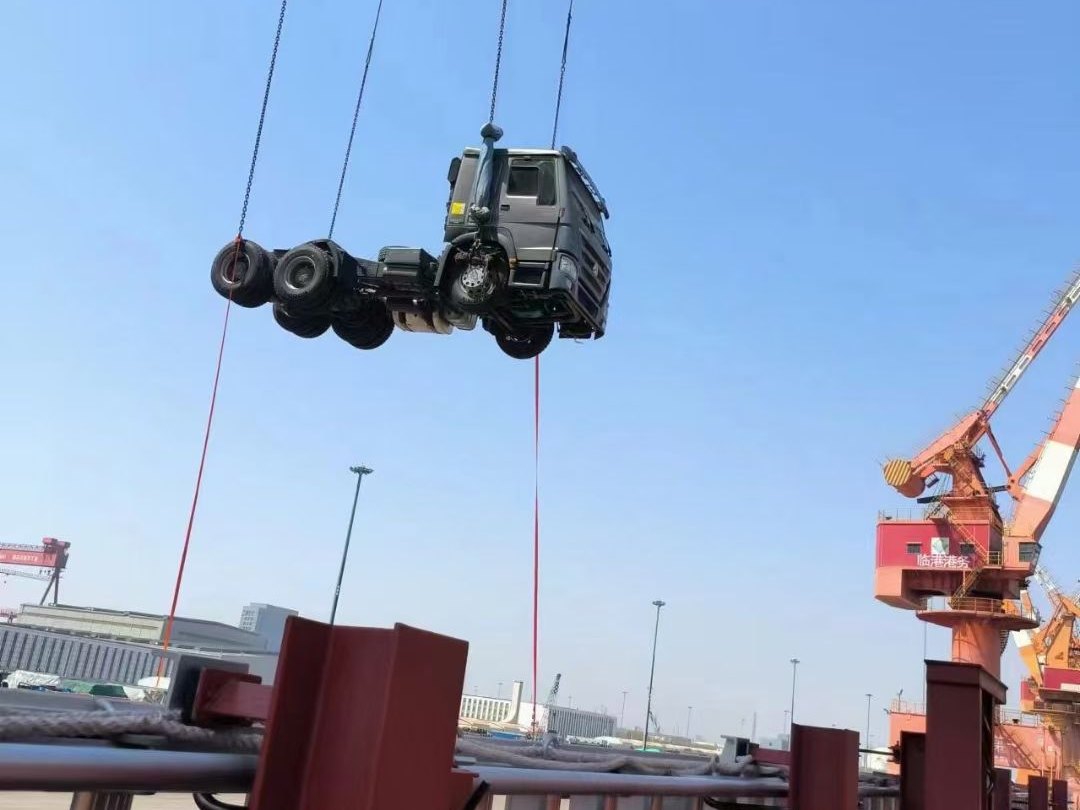 A truck chassis lifted by a quay crane at the loading port, gantry cranes in the background