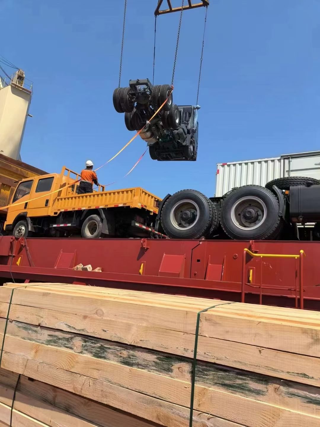 A worker guiding a tractor unit being lowered onto a flatbed, timber dunnage stacked in the foreground